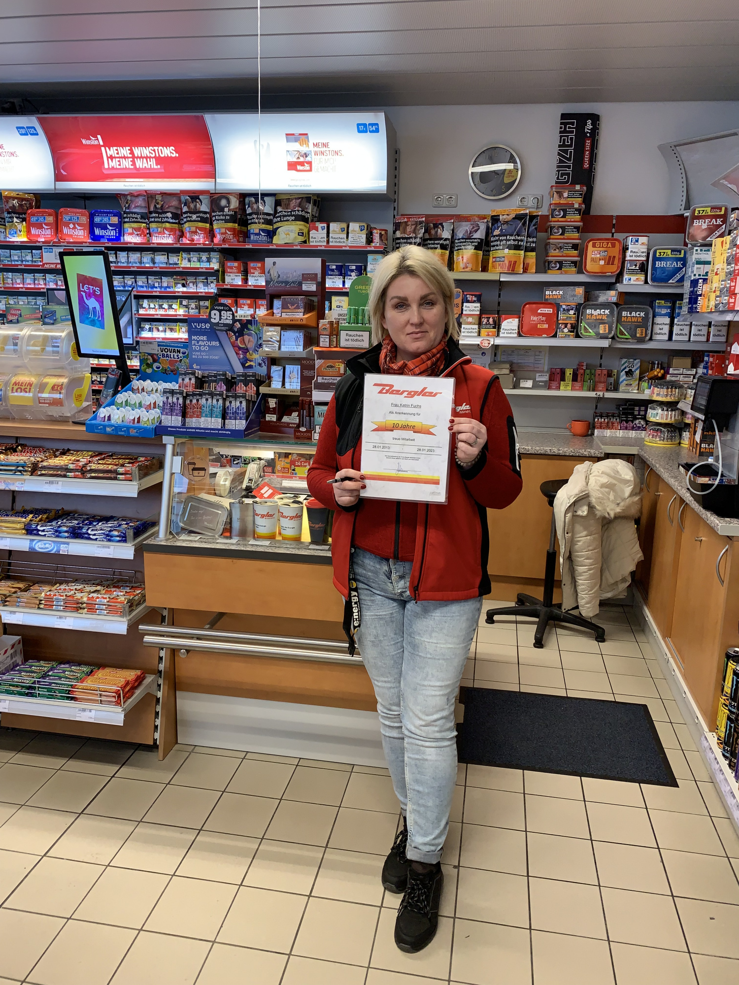Im Hintergrund der Kassenbereich mit einem Teil des Shop-Sortiments der Tankstelle Bergler in Weiden. Im Vordergrund die Jubilarin mit Urkunde für die lange Betriebszugehörigkeit. 