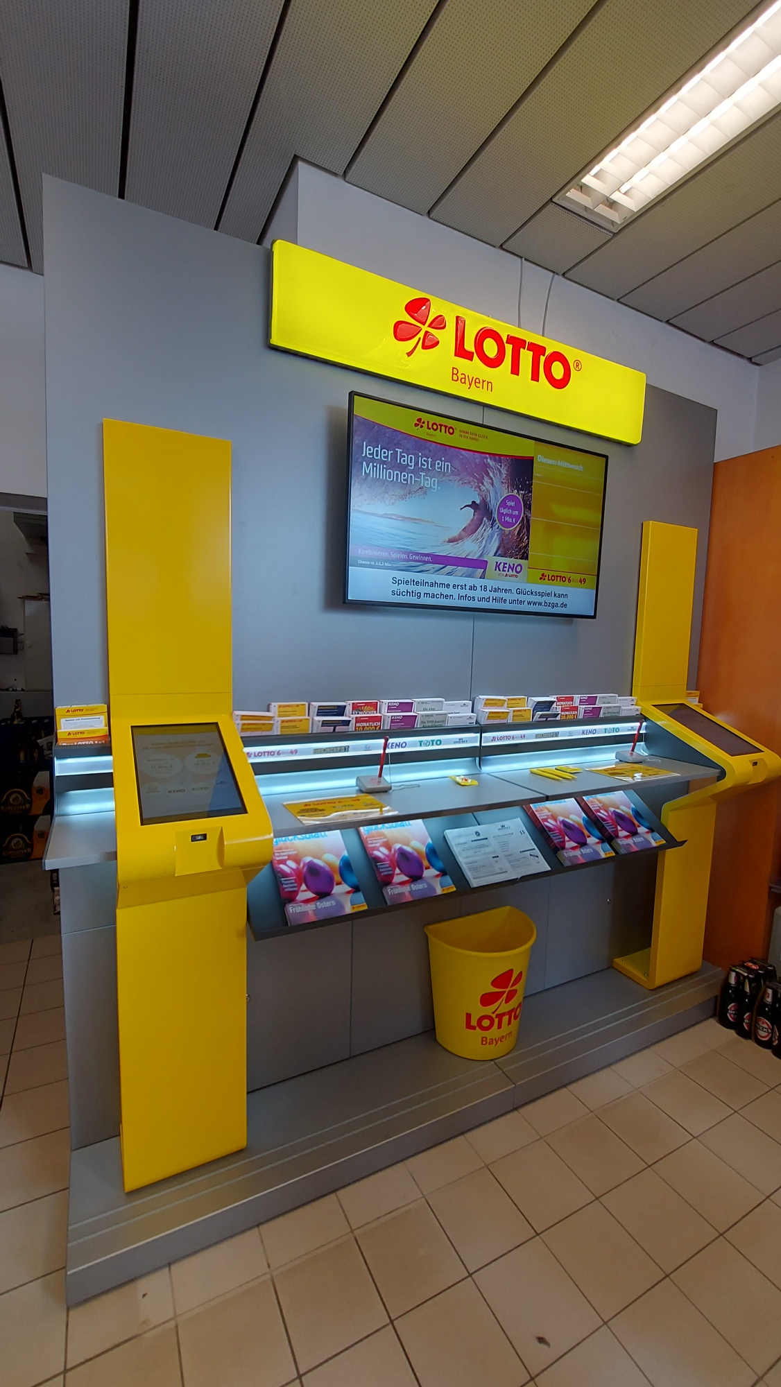 Lotto Annahmestelle in Weiden. An der Lottowand hängt mittig oben die Leuchtreklame "Lotto" mit roter Schrift auf gelbem Hintergerund, darunter ein größerer Bildschirm. Links und rechts in Gelb sind kleinere Bildschirme eingefasst. Zwischen dieser Einfassung die graue Ablage, auf der die Tippscheine ausgefüllt werden können.