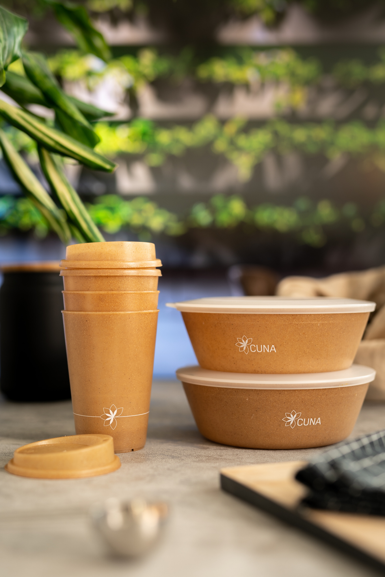 Links stehen gestapelte Mehrwegbecher. Auf den bräunlichen Bechern ist in weiß das Logo von CUNA, dem Hersteller, abgebildet. Rechts zwei gestapelte Bowls mit Deckel, ebenfalls in leicht bräunlicher Farbe mit weißem CUNA Schriftzug. Im Hintergrund erkennt man unscharf grüne Pflanzen.